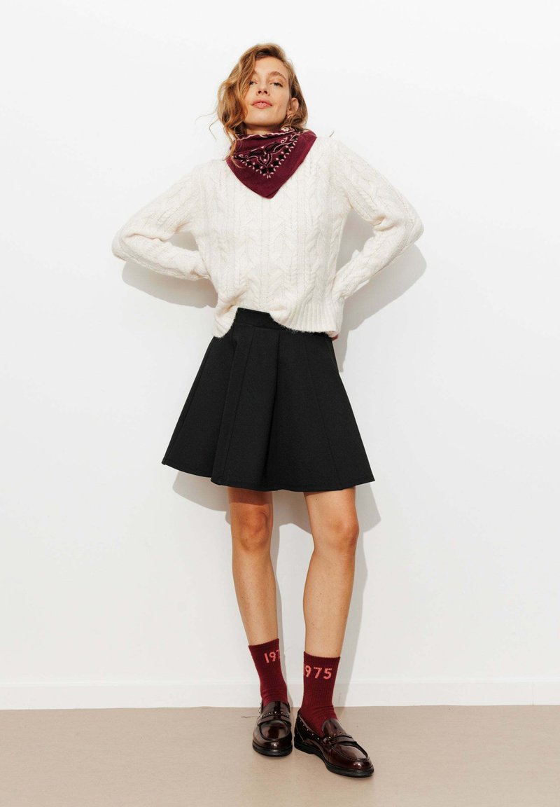 Pull en tricot blanc avec un motif en cablage, jupe noire évasée, écharpe bordeaux, et chaussettes assorties avec le texte "1975", accompagnées de chaussures sombres.