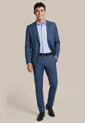 Junger Mann lächelt, trägt einen blauen Anzug mit hellblauem Hemd, schwarzem Gürtel und schwarzen eleganten Schuhen, steht vor einem schlichten Hintergrund.