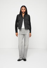 Ženska v črnem napihnjenem telovniku, črnem dolgih rokavih, svetlo sivih kavbojkah, črnih čevljih in majhni črni torbici, stoji na beli ozadju.