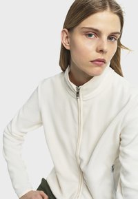 Veste en polaire blanche avec un col montant, fermeture éclair intégrale et poches latérales. Texture lisse avec des coutures discrètes et un design minimaliste.