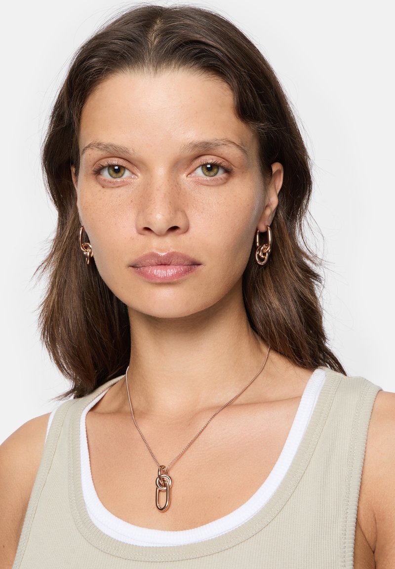 Smykker i rosaguld består af en kædehalskæde med et snoet design og matchende hooopøreringe. Modellen har en beige top med hvidt kant.