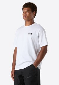 Ung mand med kort krøllet hår iført en hvid The North Face t-shirt og sorte bukser, med den ene hånd i lommen, vendt mod venstre.