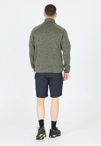Olivgrön fleecejacka med hög krage, kombinerad med mörka shorts. Anmärkningsvärda detaljer inkluderar textil med struktur och dragkedja i halsen.