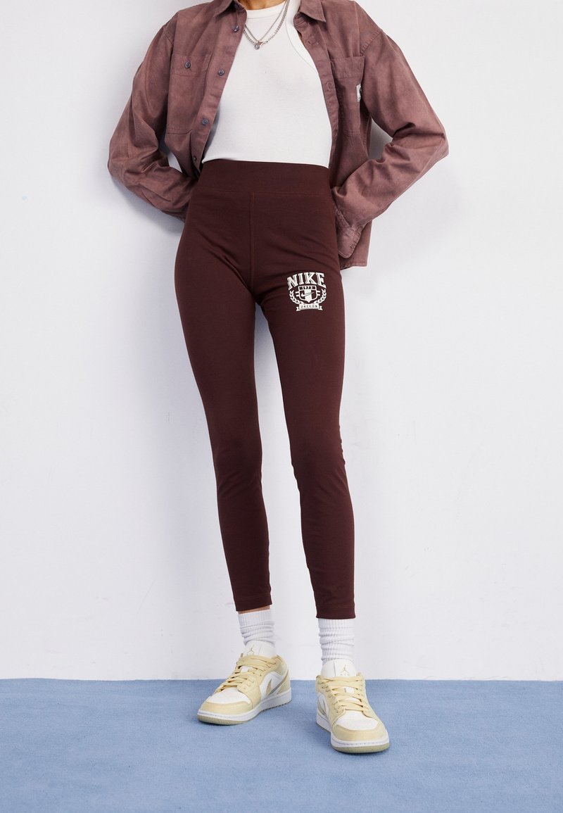 Hnědé legíny s vysokým pasem s potiskem loga Nike na stehně, zkombinované s bílým upnutým topem a světle hnědou košilí na knoflíky, a žlutými teniskami.