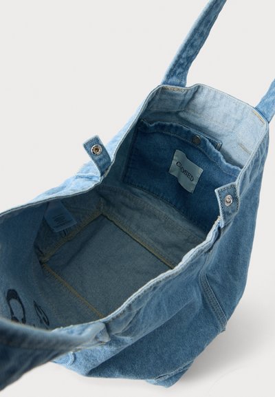 Sac cabas en denim bleu clair avec fermeture à bouton-pression et poche intérieure étiquetée "CLOSED", présenté ouvert et vide sur un fond neutre.