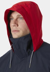 Marineblauwe hooded jacket met een rode capuchon, trekkoordverstelling en een hoge kraag, gemaakt van een gladde, waterafstotende stof.