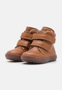 Brązowe skórzane buty z dwiema regulowanymi paskami na rzep, teksturowanymi gumowymi podeszwami i miękką wyściółką. Okrągły nosek i niski krój za kostkę.