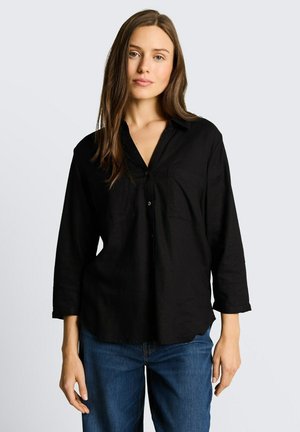 Junge Frau mit langen braunen Haaren, die ein schwarzes, knöpfbares Hemd mit Dreiviertelärmeln und blaue Jeans vor einem hellen Hintergrund trägt.