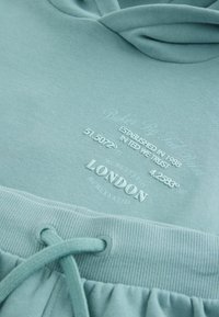 Világoskék pamut kapucnis pulóver bordás mandzsettákkal és aljjal. Fehér szöveges részletekkel, beleértve "London"-t és koordinátákat. Zsinóros záródás a kapucnin.