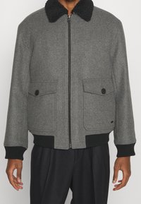 Man som bär en grå zip-up jacka med svart fleecekrage, två framfickor med knäppning och svarta ribbade muddar och fåll.