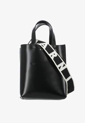 Borsa tote in pelle nera con manici corti e una larga tracolla bianca recante un testo nero in grassetto.