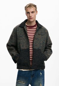 Wollmischungjacke mit Kragen, dunkler und mehrfarbiger Webstruktur, Fronttaschen und dezentem Reißverschlussdetail. Getragen über einem gestreiften Hemd.