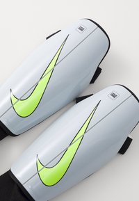 Nike scheenbeschermers van lichtgrijs plastic, met een opvallend zwart en neon groen swoosh ontwerp, voorzien van verstelbare zwarte riemen aan de achterkant.