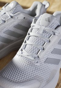 White athletic shoes featuring a textured fabric upper, gray accents, and three gray stripes. Rounded toe and flat laces complete the design.