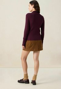 Femme portant un haut à manches longues violet, une mini-jupe en daim marron, des chaussettes beiges et des mocassins marron foncé, se tenant de dos.