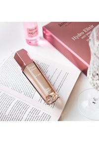Hydro Elixir mist spray med et geometrisk pink låg, gennemsigtig krop og hvid tekst, placeret på en åben bog ved siden af et glitrende glas.