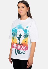 Wit katoenen T-shirt met een kleurrijke afbeelding van palmbomen en een danser-silhouet. Tekst in het roze: "Positive Vibes".