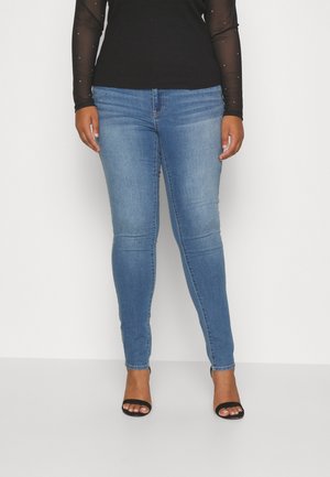 Kvinde iført lyseblå skinny jeans, sort gennemsigtig bluse med lange ærmer og sorte åbne hæle stående mod en hvid baggrund.