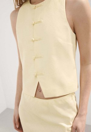 Haut sans manches jaune pâle avec cinq boucles de bouton en tissu et une fente à l'avant, porté avec une jupe taille haute assortie.