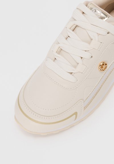 Baskets beige clair avec une tige synthétique lisse, des accents dorés et des lacets plats. Présente une forme de pied arrondi et une semelle texturée.