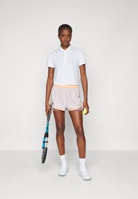 Fehér polo ing, világos rózsaszín rövidnadrág narancssárga derékrésszel és díszítéssel, fehér sportcipő, teniszkészletet és teniszbalt tart a kezében.