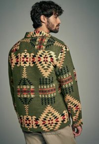 Chemise verte à motifs géométriques en rouge, beige et noir. Dotée d'un col et de poignets boutonnés, fabriquée à partir d'un tissu texturé.