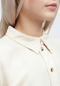 Camisa bege com colarinho, feita de um tecido texturizado, apresentando dois botões metálicos no decote e um design de colarinho clássico.