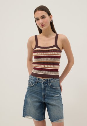 Junge Frau trägt ein gestreiftes, gestricktes Tanktop und zerrissene Jeansshorts, steht mit den Händen hinter ihrem Rücken vor einem schlichten Hintergrund.