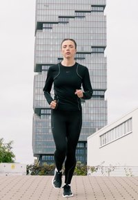 Vrouw in zwarte sportkleding die jogt op een verharde pad met een modern glazen gebouw op de achtergrond.