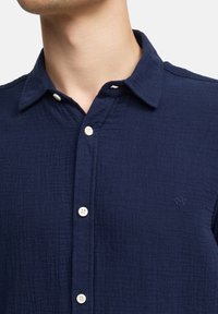 Marineblaues Hemd aus strukturiertem Baumwollstoff. Verfügt über einen kragenlosen Ausschnitt, weiße Knöpfe und ein kleines gesticktes Logo auf der Brust.