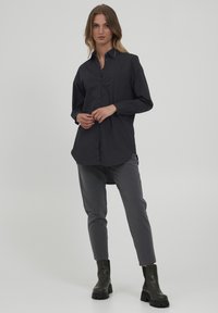 Black long-sleeve shirt with a button-down front, curved hem, and contrast collar, worn with gray tapered pants and black ankle boots.