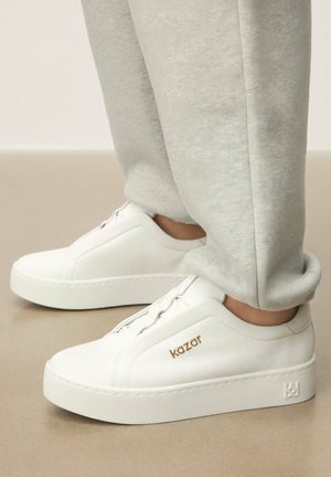 Białe skórzane sneakersy na grubej platformie, z delikatnym przeszyciem i złotym logo po bokach. W połączeniu z szarymi spodniami dresowymi.