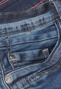 Jean en denim de couleur bleu foncé avec des coutures visibles, une petite poche avant et un détail en rivet argenté près de la taille.