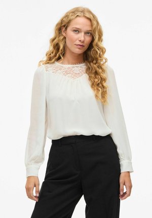 Blusa bianca con maniche lunghe, dettagli in pizzo sul colletto. Tessuto morbido e leggero, vestibilità ampia, abbinata a pantaloni neri su misura.