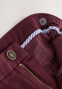 Pantalon en coton bordeaux avec une fermeture éclair en laiton, une fermeture à bouton, et un détail de ceinture rayée bleu et blanc.