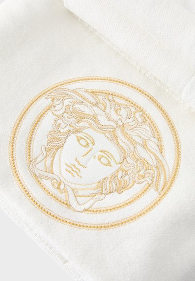 Prosop alb cu broderie aurie complexă reprezentând o față stilizată într-un design circular. Textură moale cu un finisaj elegant.
