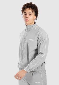 Tuta grigio chiaro realizzata in materiale liscio e leggero. Presenta dettagli bianchi, un colletto con zip e polsini e cintura elastici.