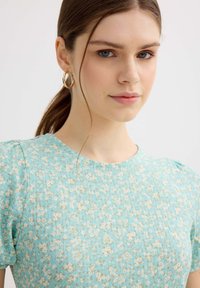 Joven mujer con ojos azules y cabello castaño en una coleta que lleva pendientes de aro dorados y un top azul claro con textura y estampado floral.