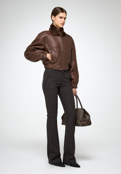 Vrouw in een bruine leren jas met bontkraag, donkergrijze flared broek, zwarte puntige schoenen, terwijl ze een gestructureerde bruine handtas vasthoudt.