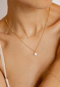 Collane multi-strato in oro con pendente in perla barocca e piccoli accenti di perle. Le catene sono delicate e con una texture liscia.