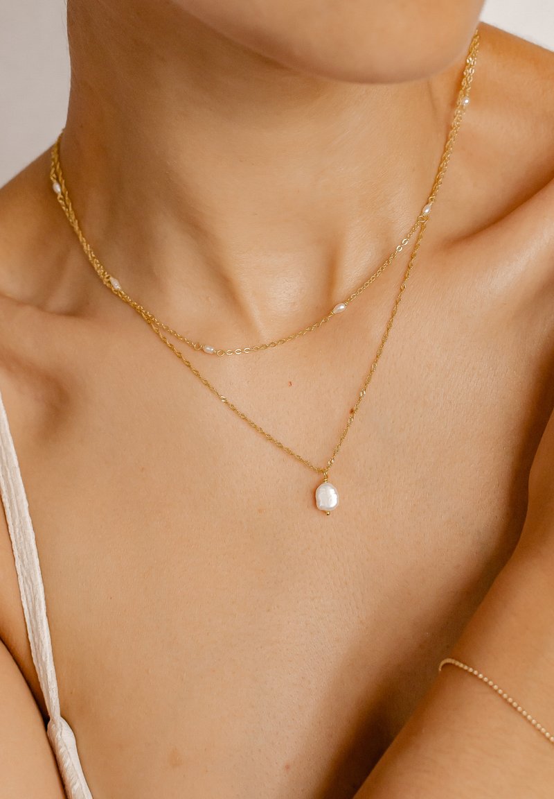 Collane multi-strato in oro con pendente in perla barocca e piccoli accenti di perle. Le catene sono delicate e con una texture liscia.