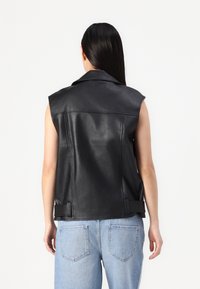 Černá kožená vesta bez rukávů s špičatým límcem, s hladkou texturou a postranními pásky. Doplněno světle modrými džínami.