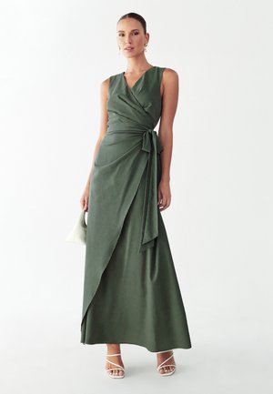 Femme portant une robe portefeuille olive sans manches avec un lien sur le côté, tenant un petit sac à main blanc et portant des talons blancs à brides.