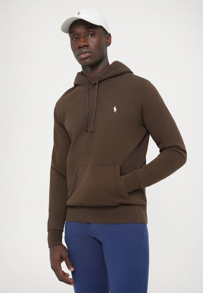 Barna kapucnis pulóver elülső zsebbel, húzózsinóros kapucnival és kis logóval díszítve. Kék nadrággal párosítva, fehér sapkát visel. Simább anyag textúrája.