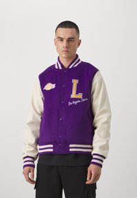 Chaqueta universitario morada con mangas blancas, logo de Los Angeles Lakers, bordado con "L" y la leyenda "Los Angeles Lakers", puños acanalados a rayas.