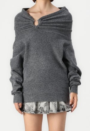 Femme portant un pull gris oversize avec un col froncé et une jupe plissée courte tie-dye sur un fond uni.