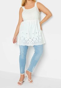 Top blanc sans manches avec un col rond, taille froncée et détails en œillets au niveau de l'ourlet, associé à un jean skinny bleu clair et des talons à lanières.