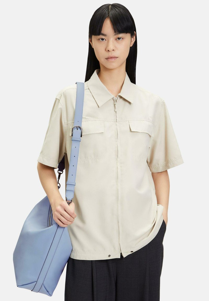 Kobieta ubrana w beżową koszulę z krótkim rękawem i kołnierzykiem oraz czarne spodnie, trzymająca dużą jasnoniebieską torbę na ramię jedną ręką.