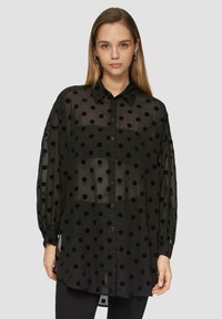 Zwarte doorschijnende blouse met zwarte stippen, knoopsluiting aan de voorkant, lange mouwen en een iets losse pasvorm. Glad textuur en lichte stof.