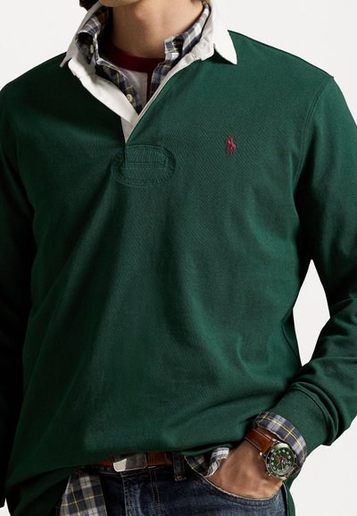 Donkergroen poloshirt met witte kraag, voorzien van een klein rood logo. Gemaakt over een geruite overhemd met knopen. Getextureerde stof, losse pasvorm.
